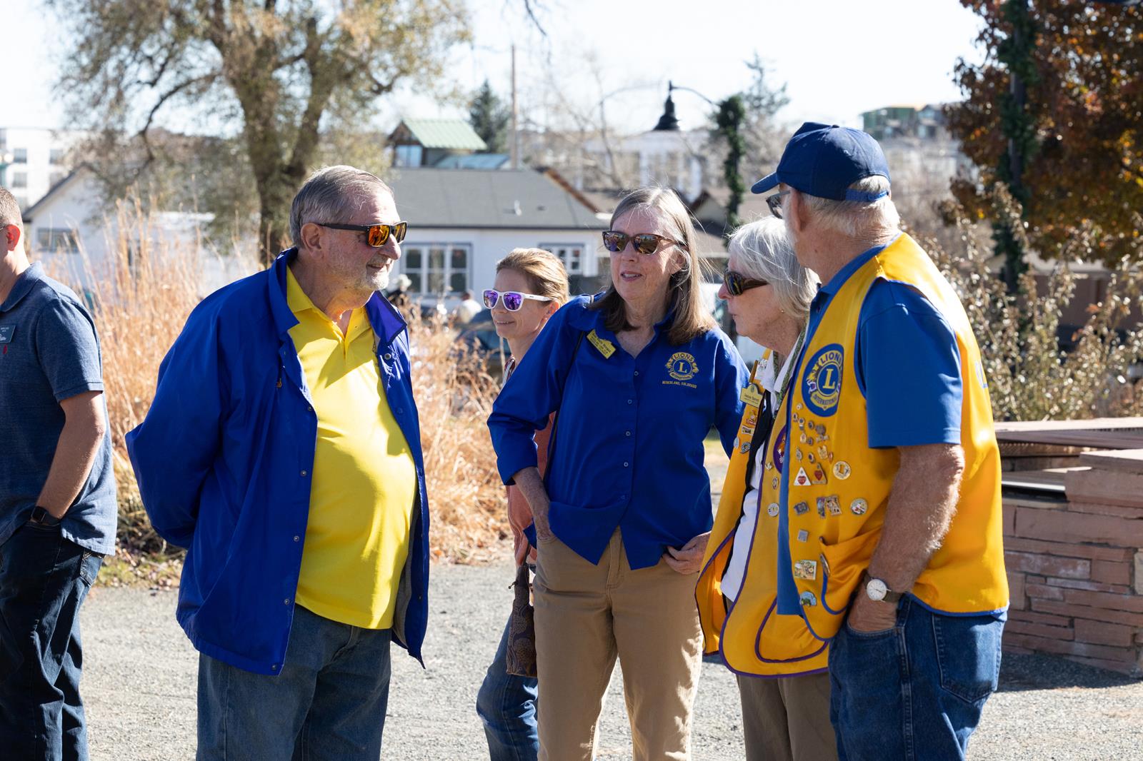 Four Lion Club representative in a semi-circle talking. Two females, two males, all wearing uniform tops of royal blue and bight yellow. A third female with long blonde hair and black shirt has back to camera in foreground.