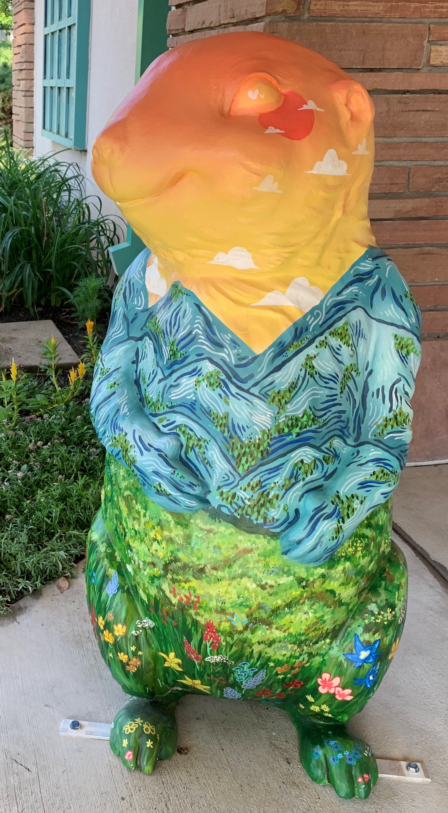 A four-foot tall 3-D fiberglass prairie dog statue painted like a landscape painting. Green grass and flowers at the feet up towards belly, blue mountains with green trees emulates a shirt and the neck and head are yellows and oranges, similar to a sunset.