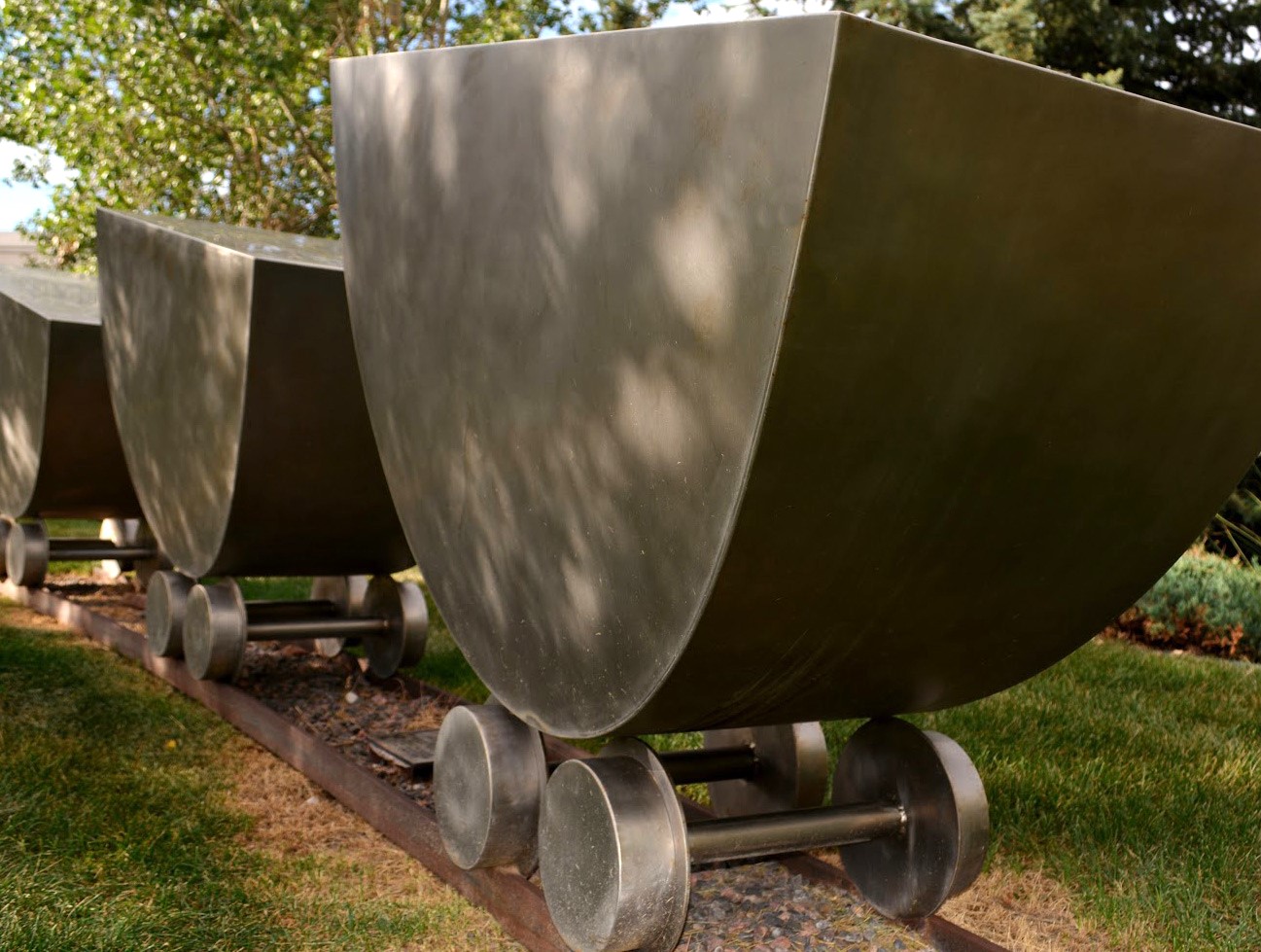 Full view of three large scale abstract coal carts, made of shiny steel. The carts sit upon a short stretch of railroad embedded in the grass.