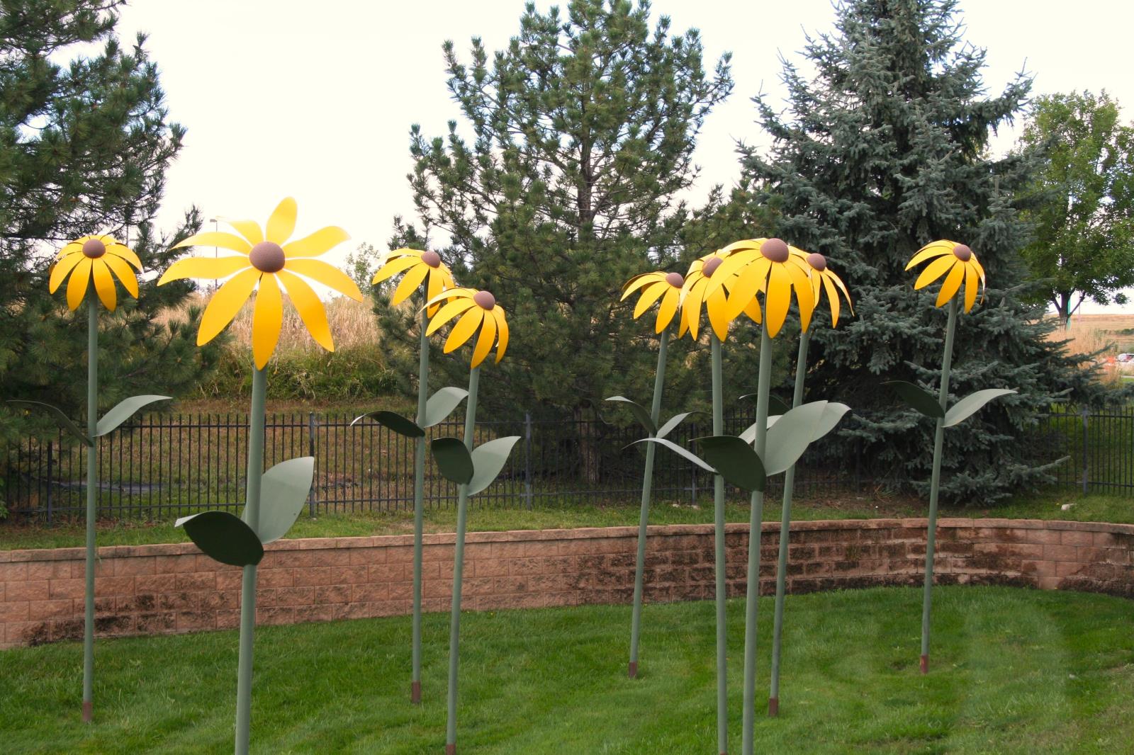 Seven larger than life metal sunflowers with green stems, yellow petals and brown centers. Large evergreen trees are behind the sunflowers.