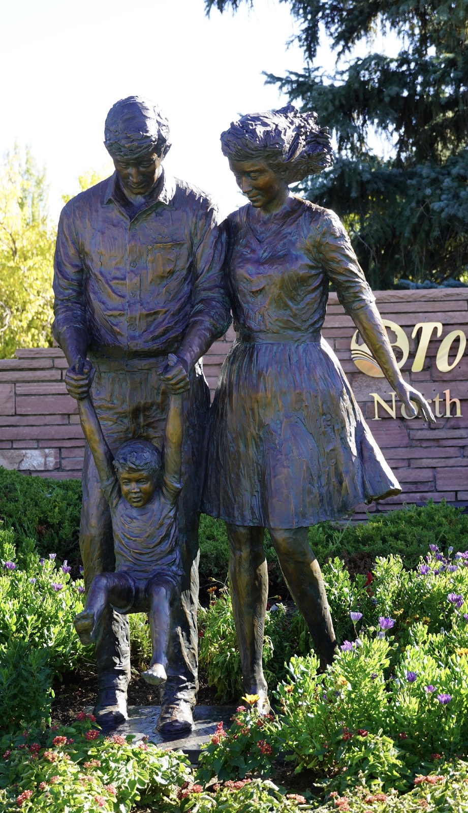 Three life-size bronze statues of two young boys and a girl all with outstretched arms as if pretending to fly. Each are barefoot and has their right leg slightly outstretched backwards.  Green grass surrounds the sculptures.