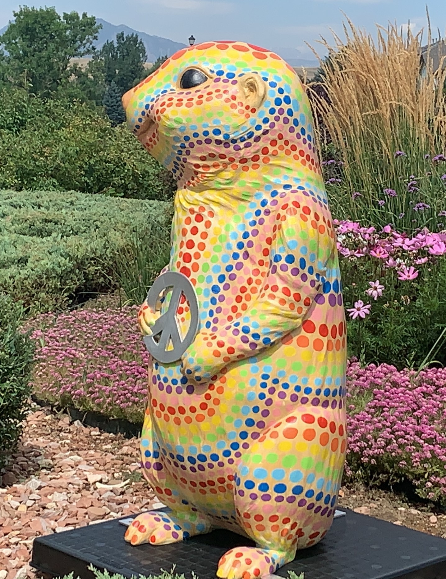 A four-foot tall 3-D fiberglass prairie dog statue painted bright yellow with a series of rainbow colored polka dots. The prairie dog is holding a large silver peace symbol.