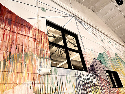 Angle view of a painted wall mural comprised of vertical lines and streaked paint that create the formation of mountain peaks in a rainbow range of colors. A black framed window, as part of the infrastructure of the concrete wall, breaks the pattern.