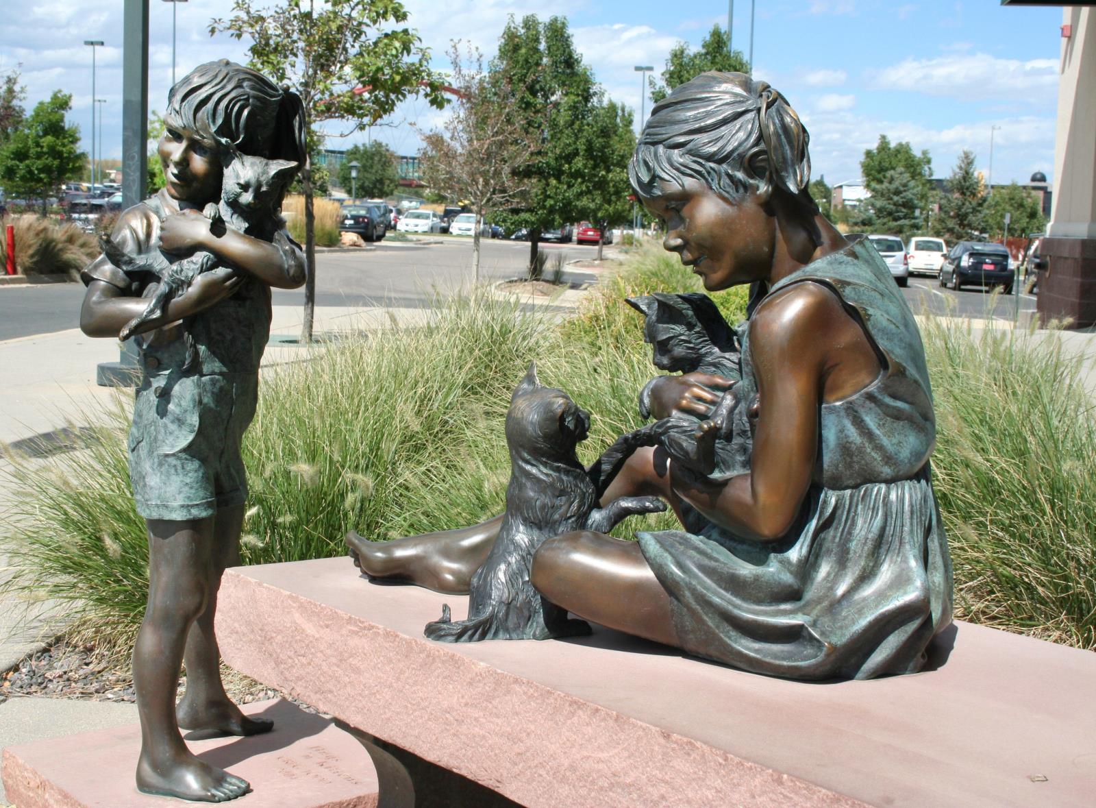 A life-size bronze sculpture of a two young girls, one standing and one seated, each holding a kitten.