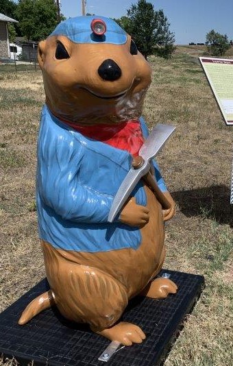 A four-foot tall 3-D fiberglass prairie dog statue with a brown painted body and fur. A blue painted coat is paired with a blue painted cap and red scarf around neck. A real flashlight is adhered to the forehead portion of the cap and the prairie dog is hold a life-size plastic pick ax.