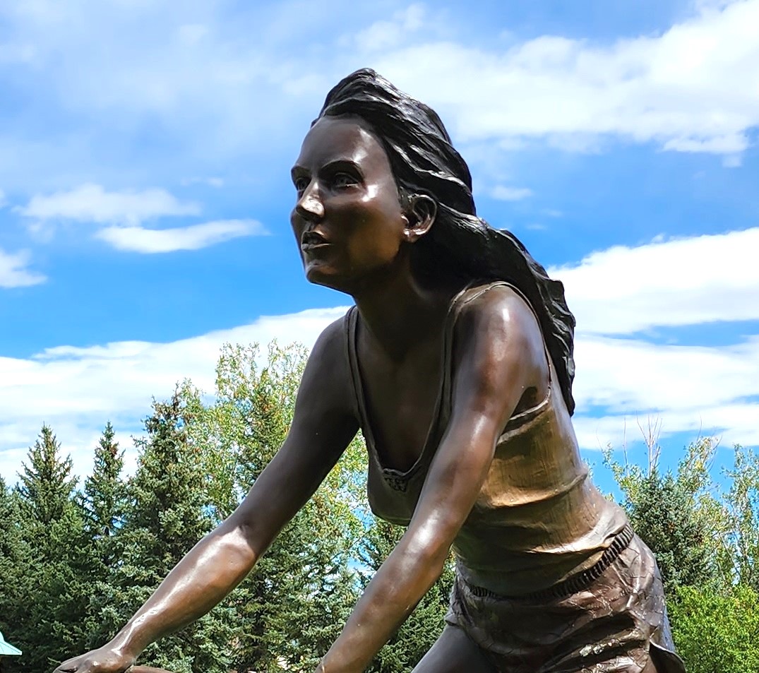 A life-size bronze sculpture, from the waist up, of a woman riding a bike wearing a tank top and below shoulder length hair flowing back from the wind.