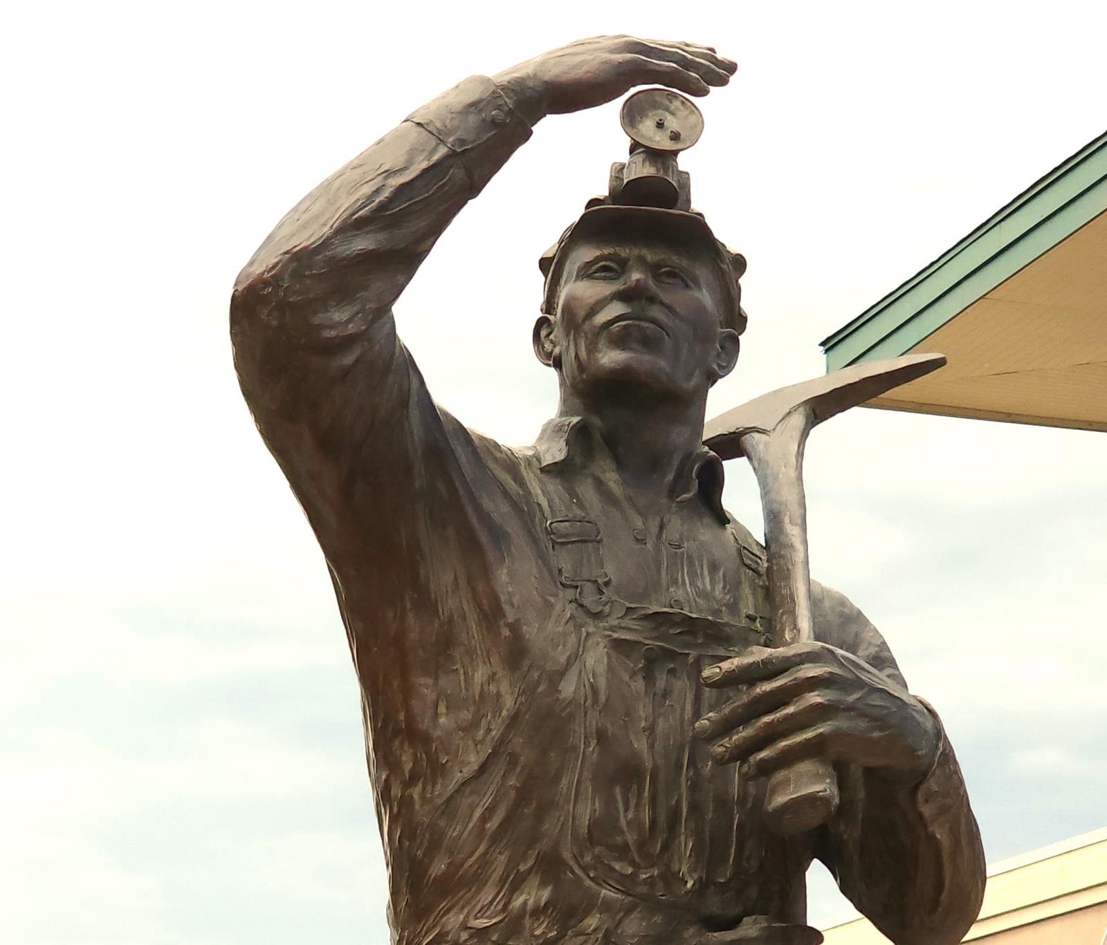 Front view of a life size bronze statue of a male miner from the waist up. He is wearing traditional coveralls, helmet with headlamp and pick ax. The miners right arm is up with his hand shielding the sun from his eyes while the left arm has the ax hoisted onto his left shoulder.