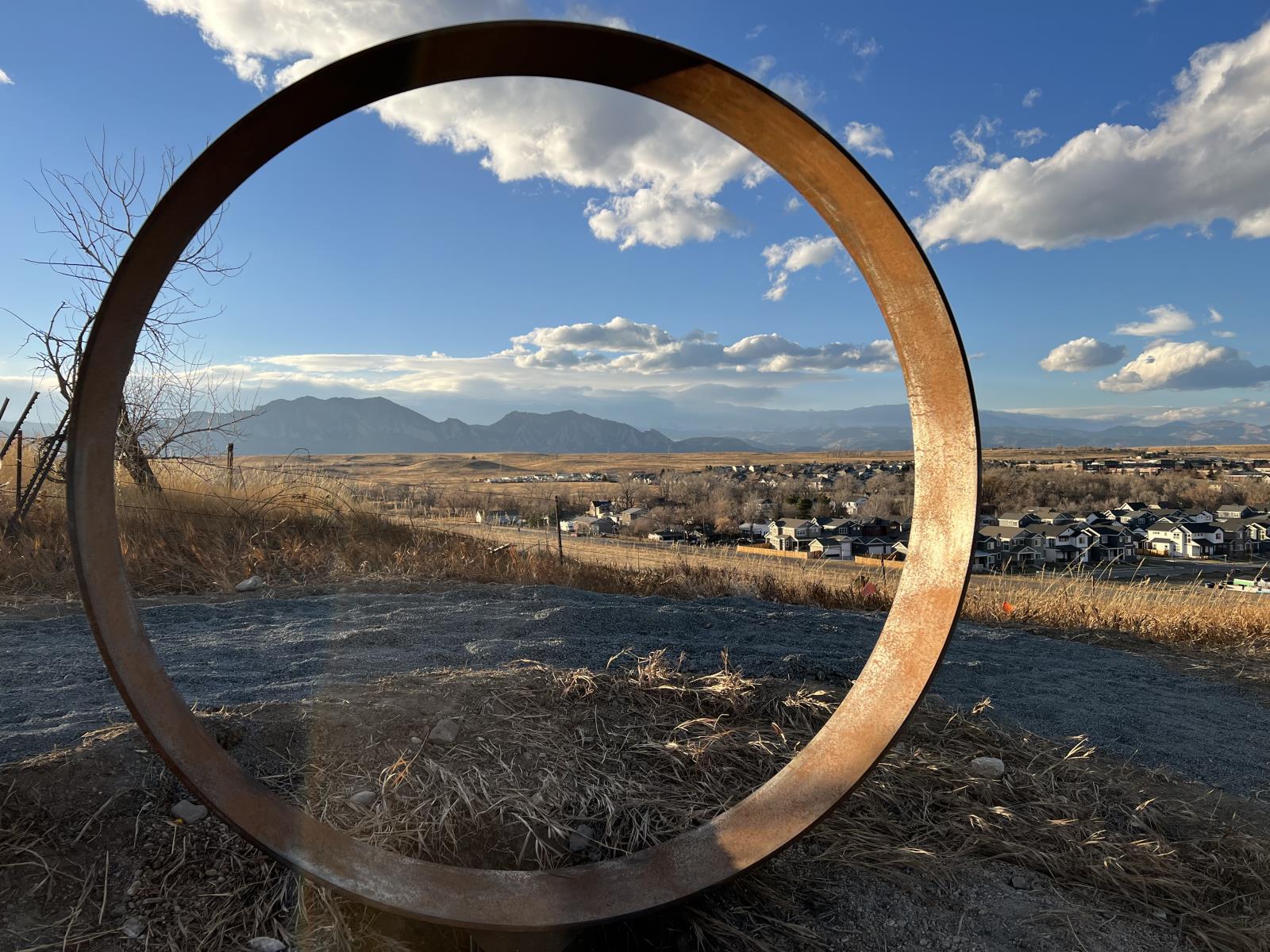 A large six-foot tall, slightly rusted steel ring installed upright in the ground. It acts like a picture frame with rolling natural grass hills, a blue sky with white clouds and the mountains beyond the sculpture.