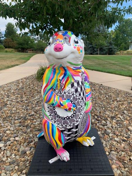 A four-foot tall 3-D fiberglass prairie dog statue  painted white with an array of geometric lines and shapes in a rainbow color palette. The colorful pattern is interrupted on its belly to reveal a white background and black lined mandala pattern.