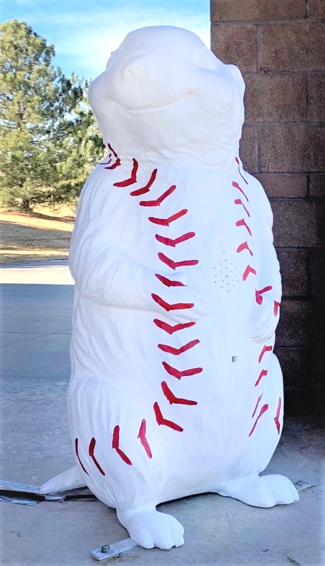 A four-foot tall 3-D fiberglass prairie dog statue  painted white with red markings from each shoulder. The markings trail down the front sides and around to the back of the body lower down. The red markings resemble the stitched on a baseball.