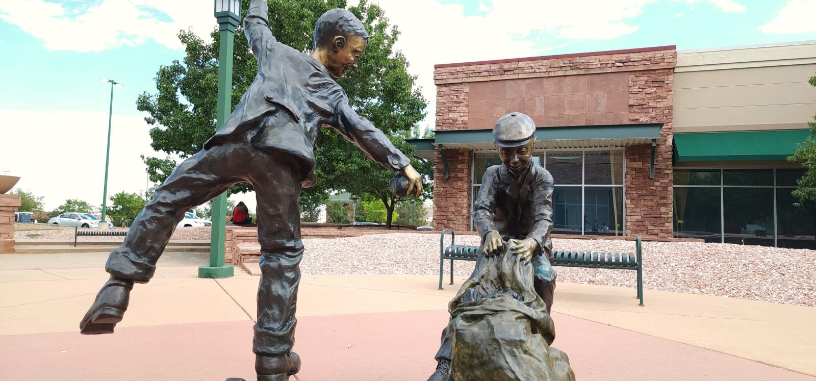 Frog-eye view of a life size bronze sculpture of two boys playfully doing their chores of gathering fallen coal chunks near the railroad tracks. One boy is balancing on one foot, coal in one hand, the other raised to help with balance. The second boy is dragging and lugging a heavy bag of coal.