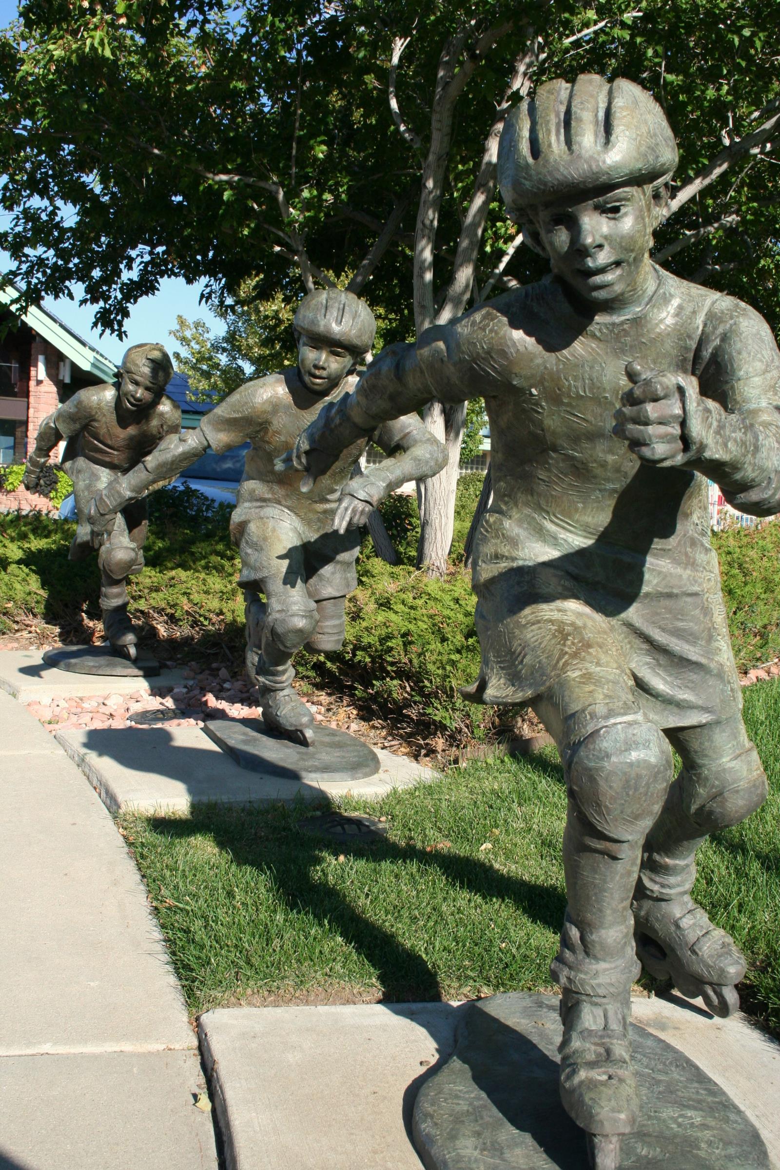 Bronze in-action statue of a young girl wearing rollerblading gear and skating with two similar boy statues behind her. Her right arm is extended back right leg and left arm are forward to indicate the skating.