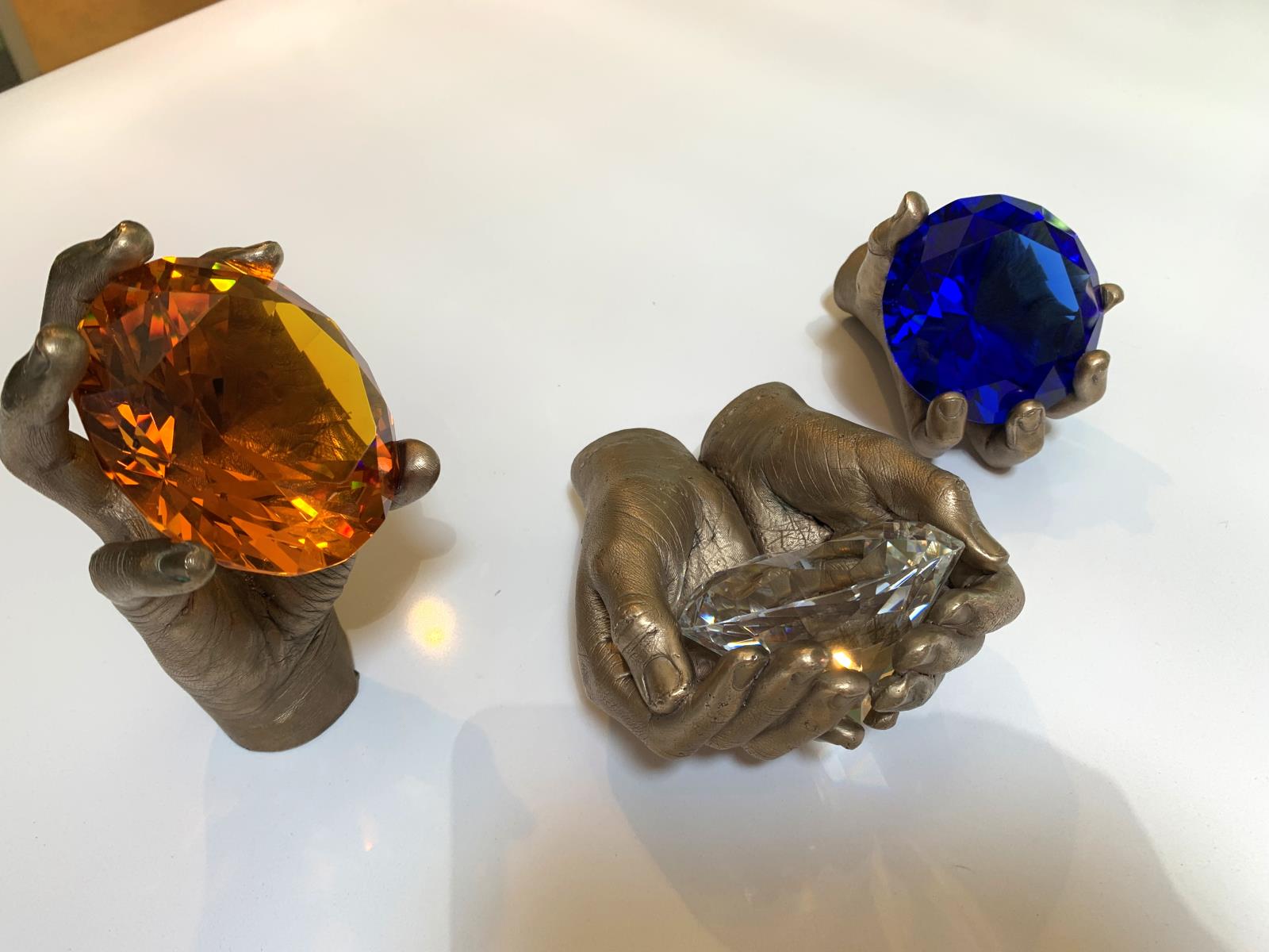 Three different bronze cast hands holding large diamond shaped gems in orange, clear and blue