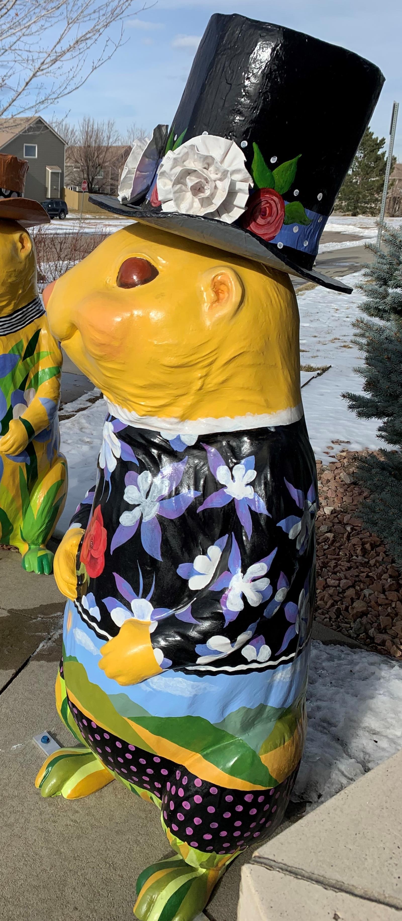 A four-foot tall 3-D fiberglass prairie dog statue painted yellow and wearing a black painted jacket with purple columbine flowers. The pants are painted in a landscape motif with green mountains and blue sky. A real paper mache black top hat is worn with large white flowers.