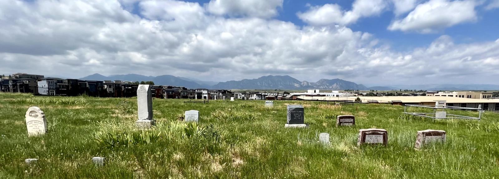 Green grass covers the Historic Cemetery site with small tombstones lining the ground and the Flatirons scraping the horizon.